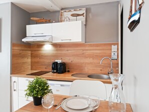 A compact kitchen with wooden cabinetry, a built-in coffee maker, a sink, and a dining area with a table set for two. - Le studio de la Ferté - In the heart of Saint Valery's main street (Saint-Valery-sur-Somme)