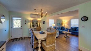 A dining area with a white table, wicker chairs, and a chandelier. There is a blue sofa and a lamp in the living area.
