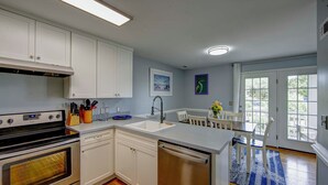 A kitchen with white cabinets, a stove, a dishwasher, a sink, and a dining area with chairs and a table. - Saltwater Therapy - Ocracoke Island (Ocracoke)