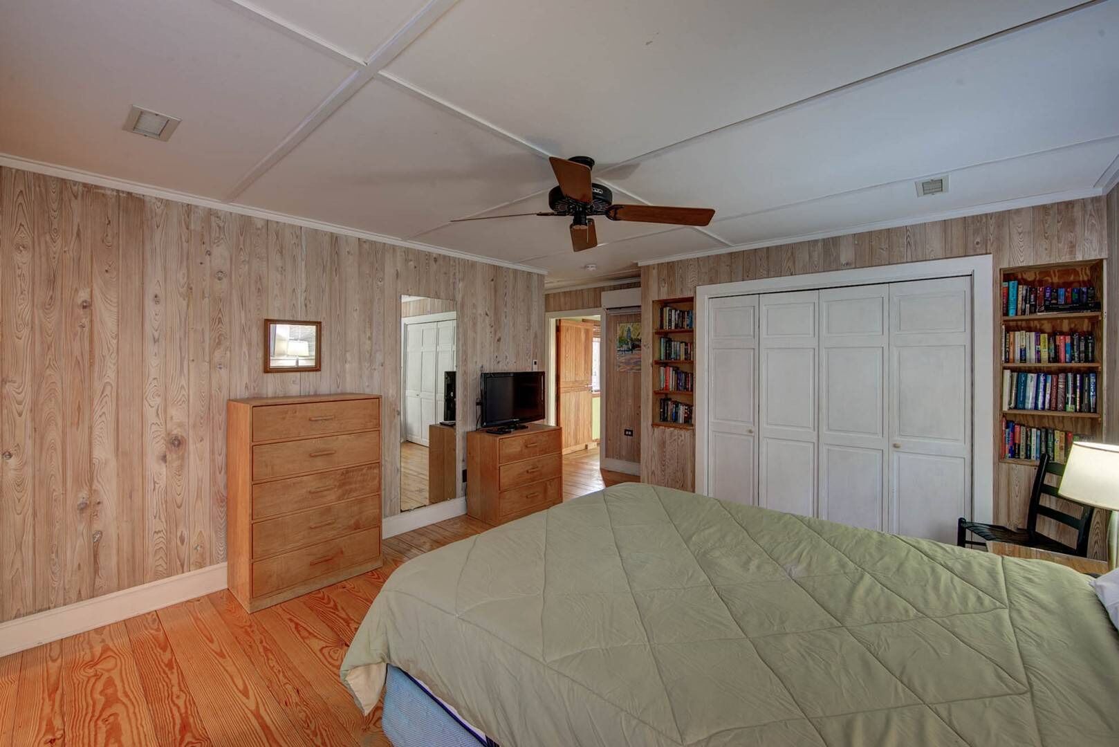 A bedroom with wooden paneling, a ceiling fan, a bed with a green quilt, a dresser, a television, a bookshelf, and a door.