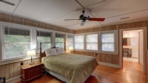 Ein Schlafzimmer mit einem Bett, Holzboden, Deckenventilator und großen Fenstern mit Jalousien.