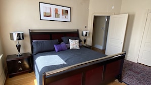 Un dormitorio con una cama de madera, dos mesitas de noche con lámparas, un ventilador de techo, un cuadro en la pared y una puerta al fondo.