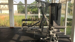Un gimnasio con máquinas de pesas y una banca, ventanas amplias y vistas al exterior con vegetación y edificios.