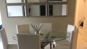 Un comedor con una mesa de vidrio rodeada por cuatro sillas blancas, un florero con ramas metálicas y un espejo colgado en la pared.