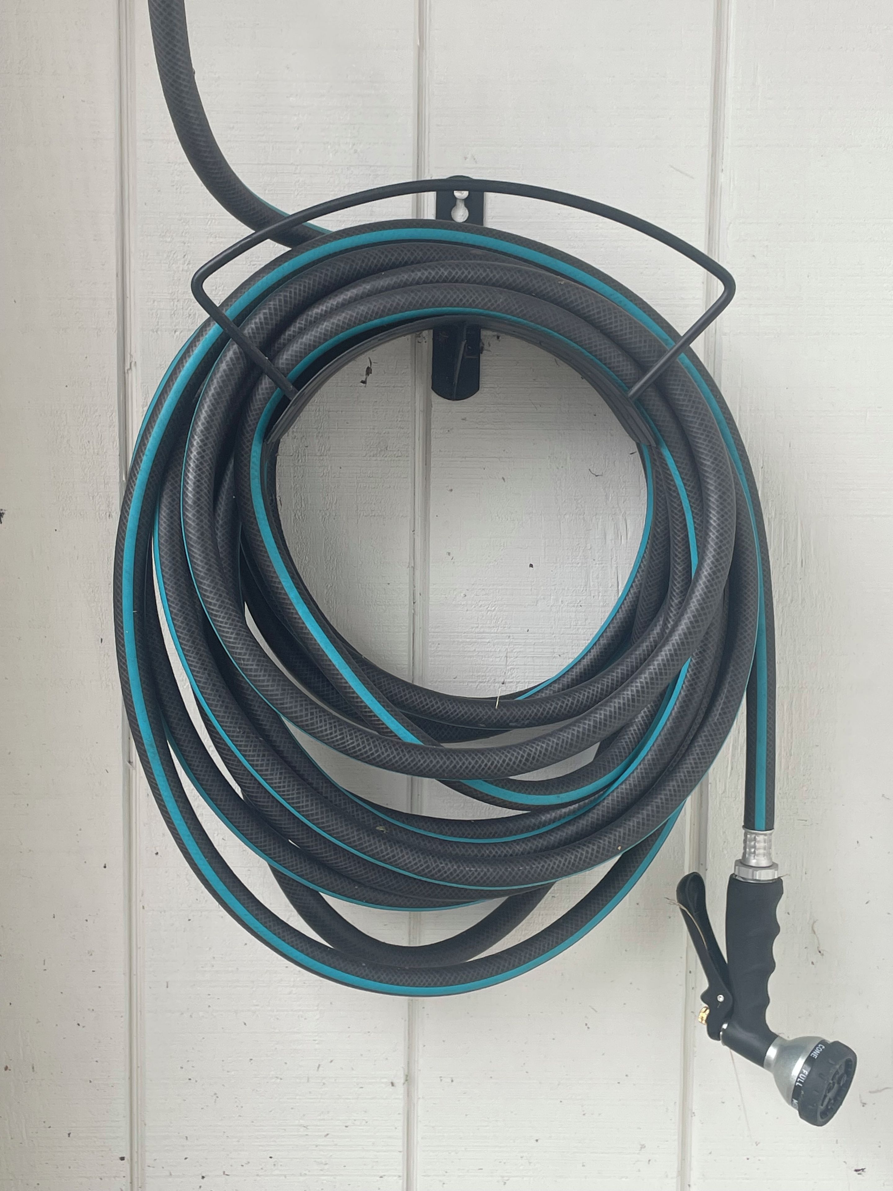 Ein aufgerollter Gartenschlauch mit Schwarz-Türkis-Farben, verbunden mit einem schwarzen Wasserhahn mit silberner Düse, vor einer weißen Wand.