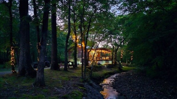 A rustic cabin surrounded by tall trees with string lights, a small stream, and a well-lit interior.