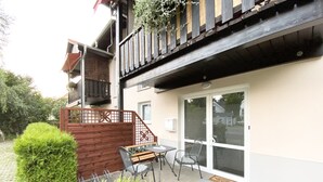 An outdoor patio with a table and chairs, a wooden fence, and a balcony with hanging plants.