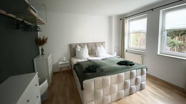 A modern bedroom with a large bed, a bedside table, a dresser, and a window with blinds.