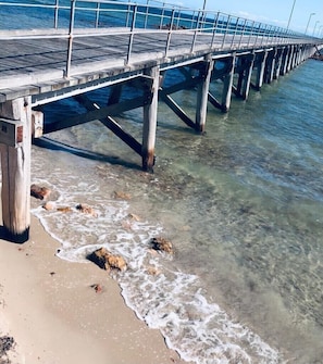 Um cais se estendendo sobre águas claras e rasas, com uma praia de areia ao final.