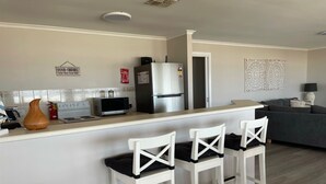A modern kitchen with a white countertop, a microwave, a refrigerator, and a coffee machine. There are three white bar stools with black cushions. A decorative vase is on the counter, and a sign reads "GOOD FRIENDS." - Stunning beachfront view ,  (North Beach)