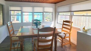 Uma área de refeições com mesa de madeira e quatro cadeiras, janelas amplas com persianas e vista para o exterior.