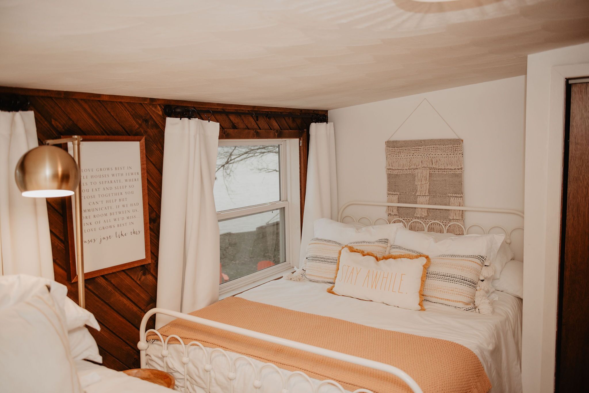 Ein gemütliches Schlafzimmer mit Bett, Nachttischlampe, einem gerahmten Zitat an der Wand und Blick aus dem Fenster.