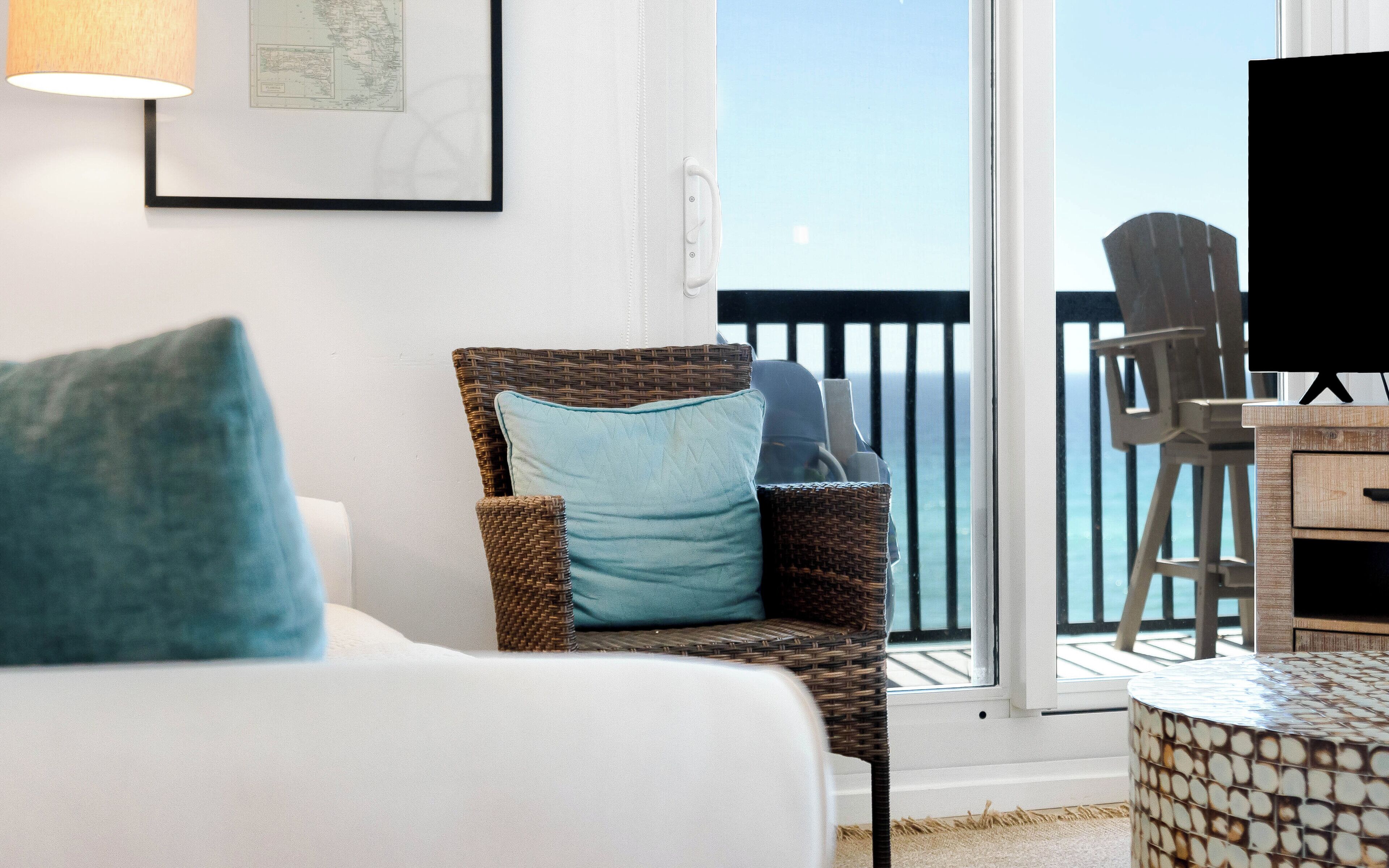 A wicker chair with a cushion, a white sofa, a framed map, and a view of the ocean through a balcony door.