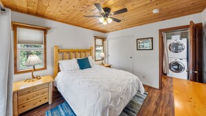 A bedroom with a wooden ceiling, a bed with white bedding, a nightstand with a lamp, a window with blinds, and a washing machine in the corner. - Mountain View: Gorgeous Views, AC, Jacuzzi bathtub! (Idyllwild)