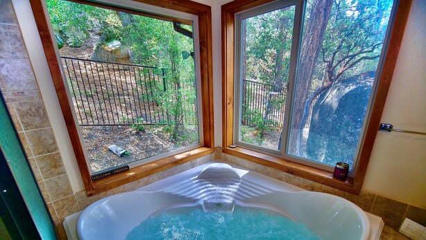 A bathroom with a large bathtub, a window with a view of a backyard, and a wooden bench. - Mountain View: Gorgeous Views, AC, Jacuzzi bathtub! (Idyllwild)