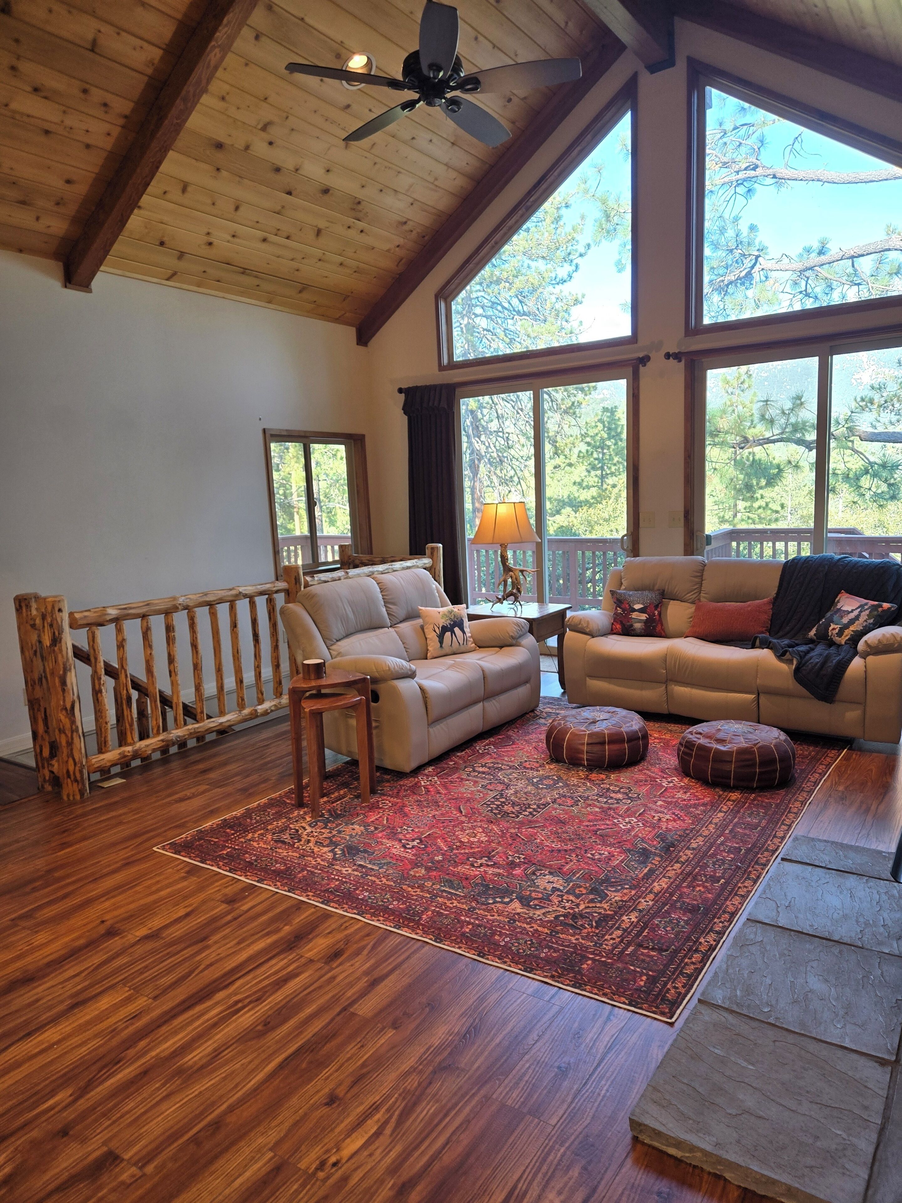 A spacious living room with a wooden ceiling, large windows, a red patterned rug, and two sofas.