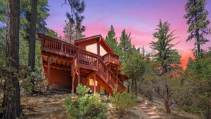 A two-story wooden cabin with a balcony, surrounded by trees and a dirt path leading to the entrance. - Mountain View: Gorgeous Views, AC, Jacuzzi bathtub! (Idyllwild)