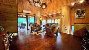 A spacious living room with wooden floors, a sofa set, a coffee table, and a dining table. There is a large window providing natural light and a view of the outdoors. - Kruger Kumba - Mugwenzi (Marloth Park)