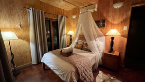 A wooden-paneled bedroom with a bed, two bedside lamps, a nightstand, and a window with curtains. - Kruger Kumba - Mugwenzi (Marloth Park)