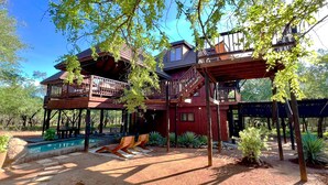 A wooden cabin with a pool, deck chairs, and a patio area surrounded by trees. - Kruger Kumba - Mugwenzi (Marloth Park)