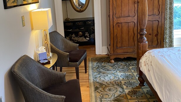 Une chambre d’hôtel avec un lit, deux fauteuils, une commode, un miroir et un tableau au mur.