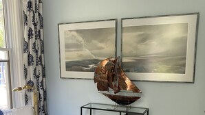 Un salon avec un canapé blanc, une table basse en verre, un pouf en peau de vache et deux tableaux représentant des paysages, encadrés et accrochés au mur.