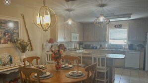 A dining area with a wooden table and chairs, a chandelier, and a kitchen in the background. - Spacious condo, fully furnished! Steps from white sugary sand of Gulf Shores! (Gulf Shores)