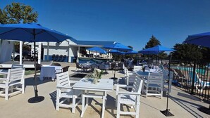 An outdoor patio area with white tables and chairs, blue umbrellas, and a pool in the background.