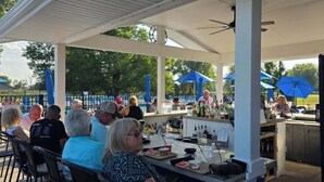 An outdoor patio area with tables and chairs, a bar with various bottles, and umbrellas providing shade.
