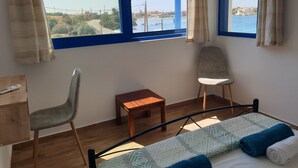 A bedroom with a bed, two chairs, a small wooden table, and large windows offering a view of the sea and buildings. - Sea View Holiday House (Archangelos)