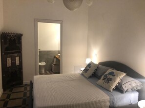 Une chambre à coucher avec un lit, une table de chevet, une lampe, une armoire et une salle de bain visible à travers une porte ouverte.