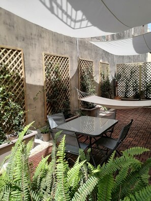 Un espace extérieur aménagé en terrasse, avec une table et des chaises en verre, entouré de plantes et d’un mur en béton.