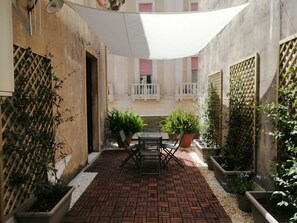 Une cour avec un sol en briques, des plantes en pot, une table avec des chaises et un auvent blanc.