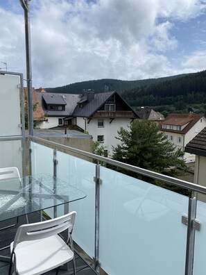 A balcony with a glass table and chairs, overlooking a residential area with houses and a forested hill. - Exclusive 2-room loft apartment in Titisee-Neustadt (Titisee-Neustadt)