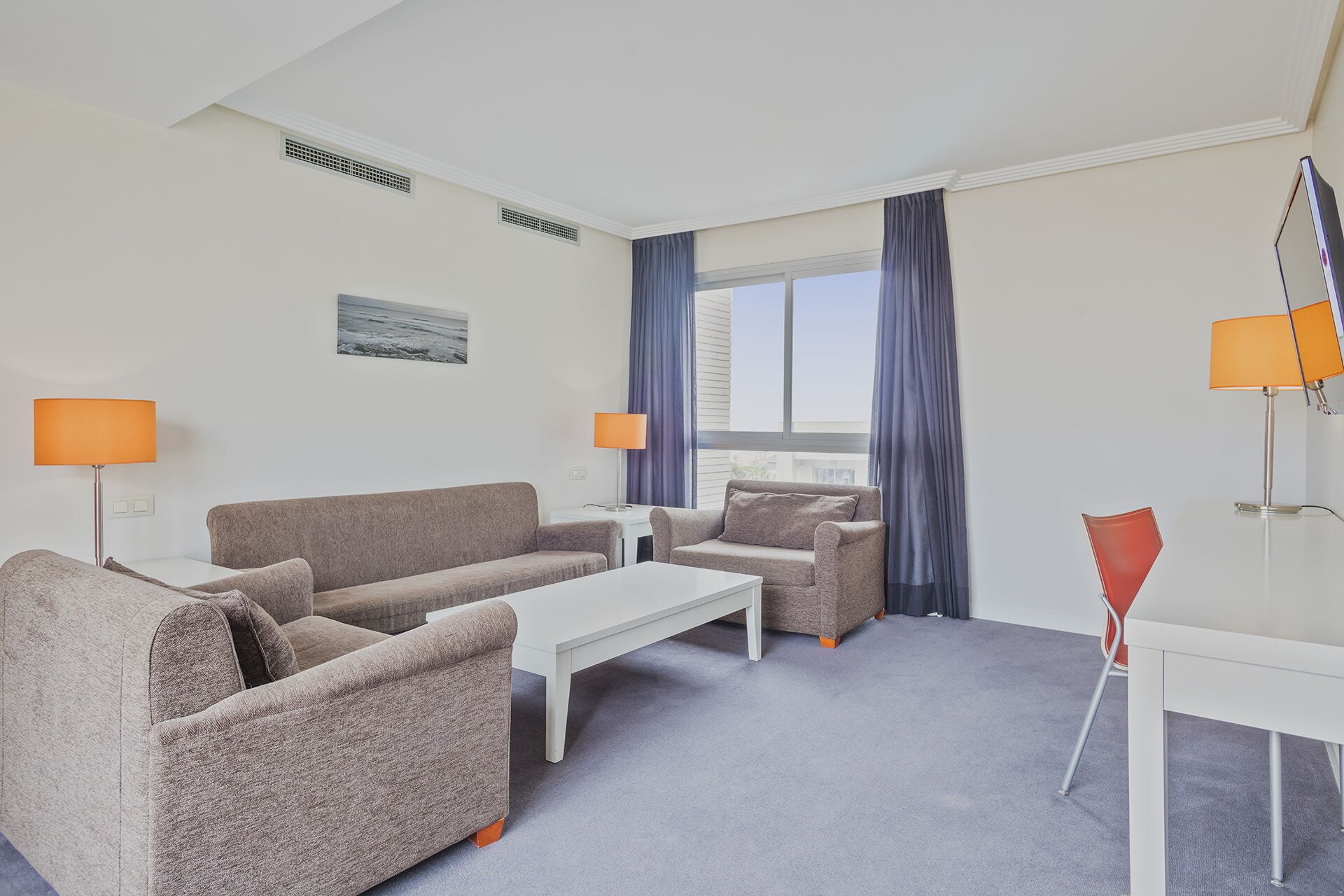 A modern hotel room with a grey sofa set, a white coffee table, and a small white desk. There are two lamps, one on each side of the room, and a view of the city through large windows with blue curtains.