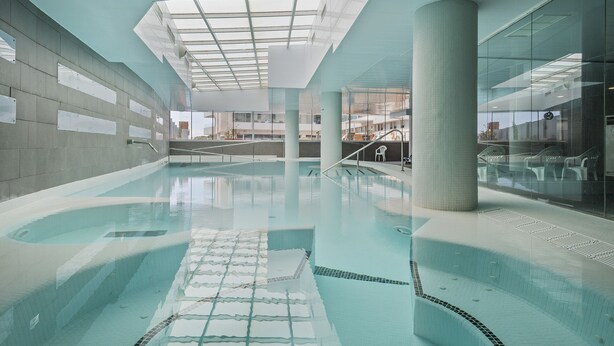 Una piscina interior con agua cristalina, techo de vidrio y vistas a los edificios exteriores.