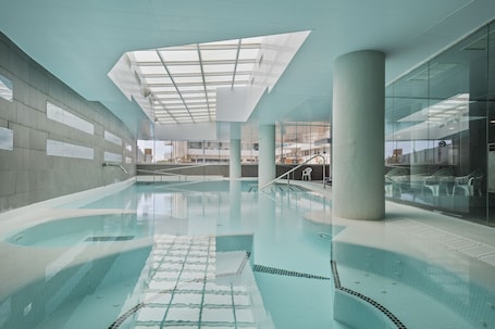 An indoor swimming pool with clear water, a glass ceiling, and a view of the outside buildings.