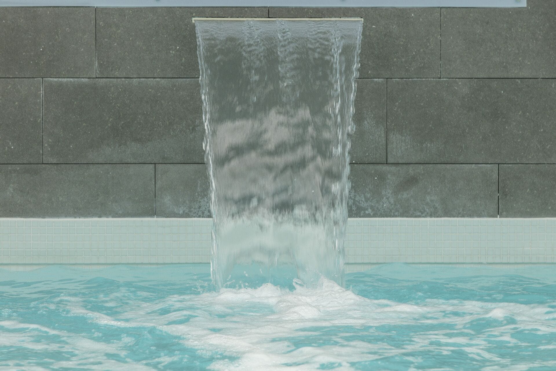 Een waterval in een zwembadgedeelte, met een betegelde muur op de achtergrond.