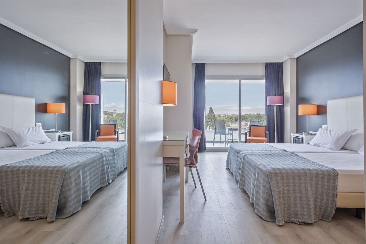 A mirrored hotel room with two beds, a desk, and a view of the outdoors.