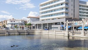 Un hotel moderno con varios pisos y balcones, vista al agua y un puente peatonal sobre una superficie acuática tranquila.
