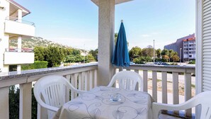 A balcony with a table and chairs, a blue umbrella, and a view of a residential area with buildings and greenery. - Holiday apartment with a view of the mountains (Baška)