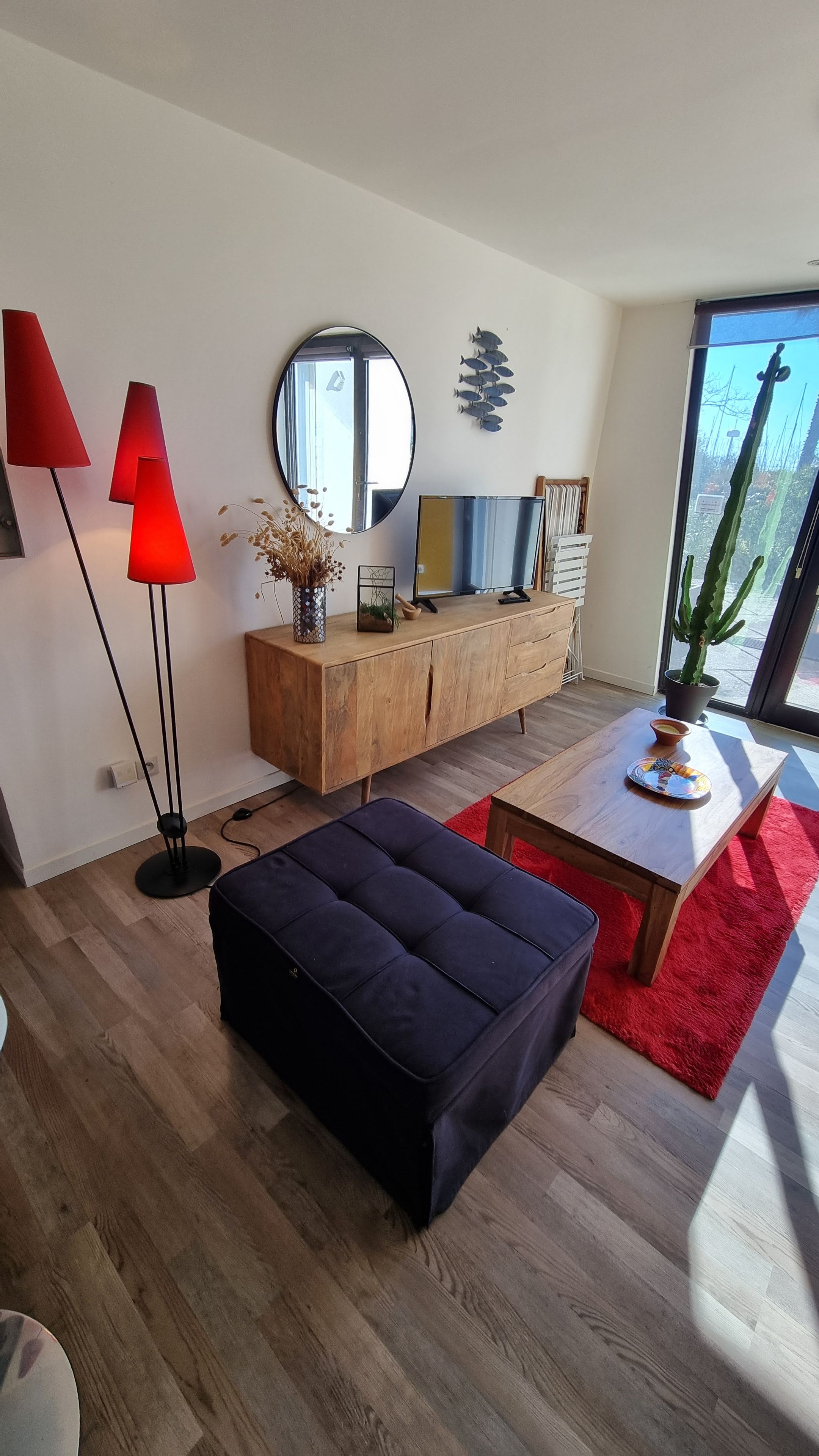A modern living room with a wooden cabinet, a round mirror, a red floor lamp, a black ottoman, and a dining table with a red rug.
