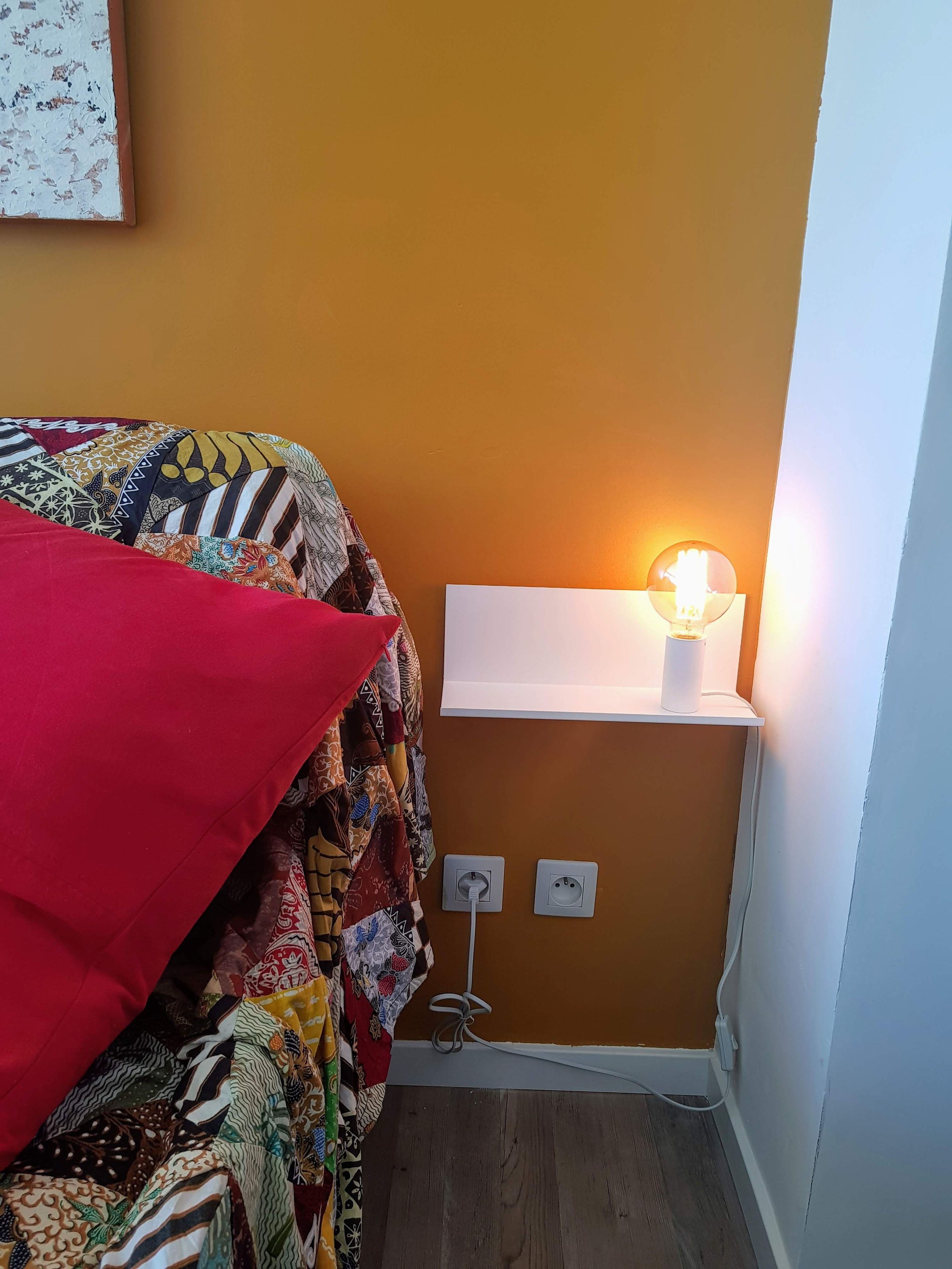 Ein Schlafzimmer mit einem Bett, das mit einer farbenfrohen Decke bezogen ist, einer Nachttischlampe und einer Steckdose.