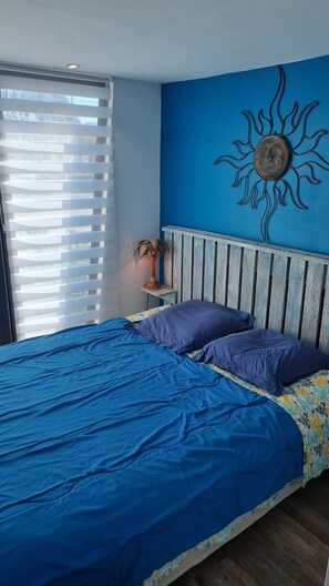 A bedroom with a bed, a wall-mounted sun decoration, a window with blinds, and a nightstand with a lamp. - Beautiful, spacious, sun-filled 47-square-meter one-bedroom apartment on one level, rated 3***, sleeps 4 (La Grande-Motte)