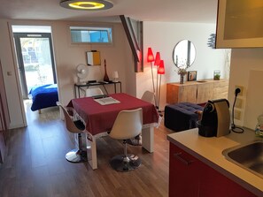 A compact hotel room with a dining area, a small kitchenette, a bed with a blue cover, a mirror, and a window with a view of the outside. - P2 spacious 47m2 sunny single-storey Classified 3*** for 4 people (La Grande-Motte)