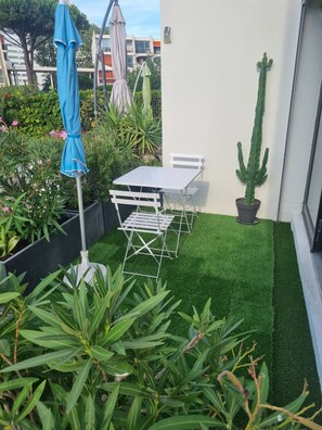 A small patio with a white table and chairs, surrounded by artificial grass, potted plants, and a cactus.
