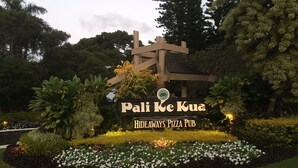 Señal de 'Pali Ke Kua Hideaways Pizza Pub' rodeada de macizos de flores bien cuidados y plantas grandes.