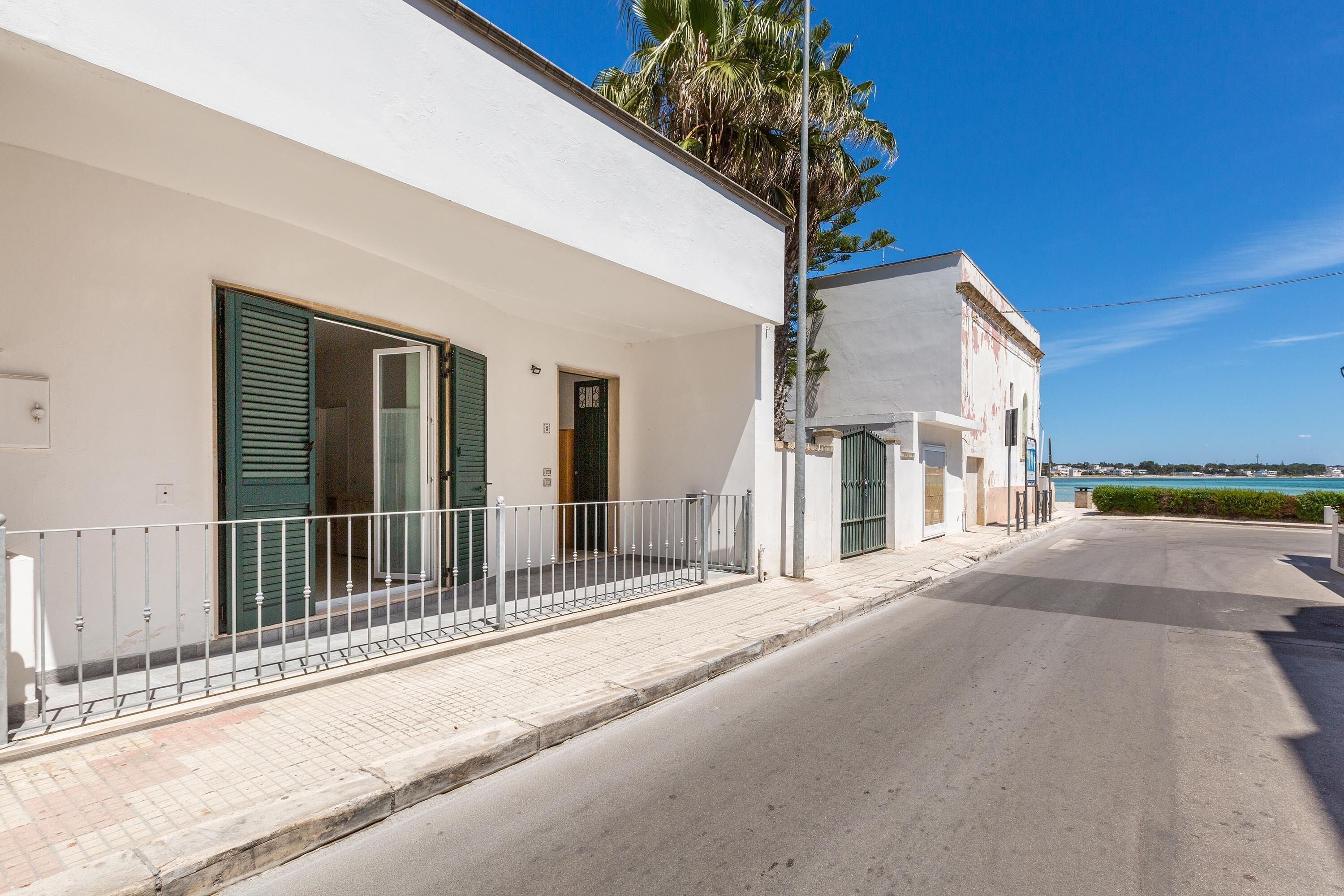 Une rue résidentielle bordée de bâtiments blancs, de volets verts et surplombée d’un ciel d’un bleu limpide.