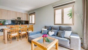 Un salon moderne avec un canapé gris, une table à manger en bois et des chaises. On y trouve également un coin cuisine avec des meubles en bois et un four à micro-ondes. Une grande fenêtre laisse entrer la lumière naturelle.