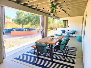 Une terrasse couverte avec une table et des chaises en bois, un tapis à rayures bleues et blanches, des plantes suspendues et une vue sur un jardin avec une piscine et du mobilier d’extérieur.